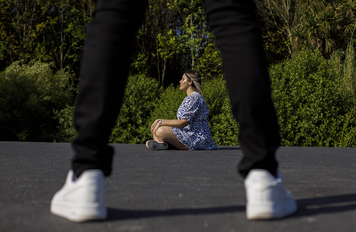 Luís de pé com pernas abertas, e ao centro, entre esse espaço, Márcia sentada, virada para o sol, num momento de reflexão.