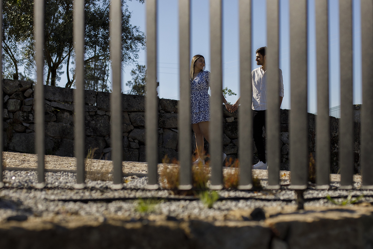 Através de uma estrutura metálica, Luís e Márcia trocam um olhar intenso, de mãos dadas, junto a um belo muro de pedra