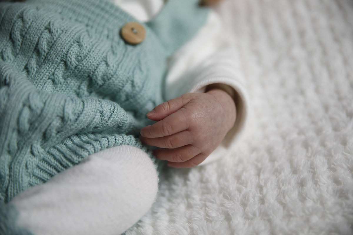 Um plano mais aproximado onde se destaca o tecido branco onde Gabriel está deitado, a sua pequena mão, e os pormenores da roupa azul e branca com botões de madeira.