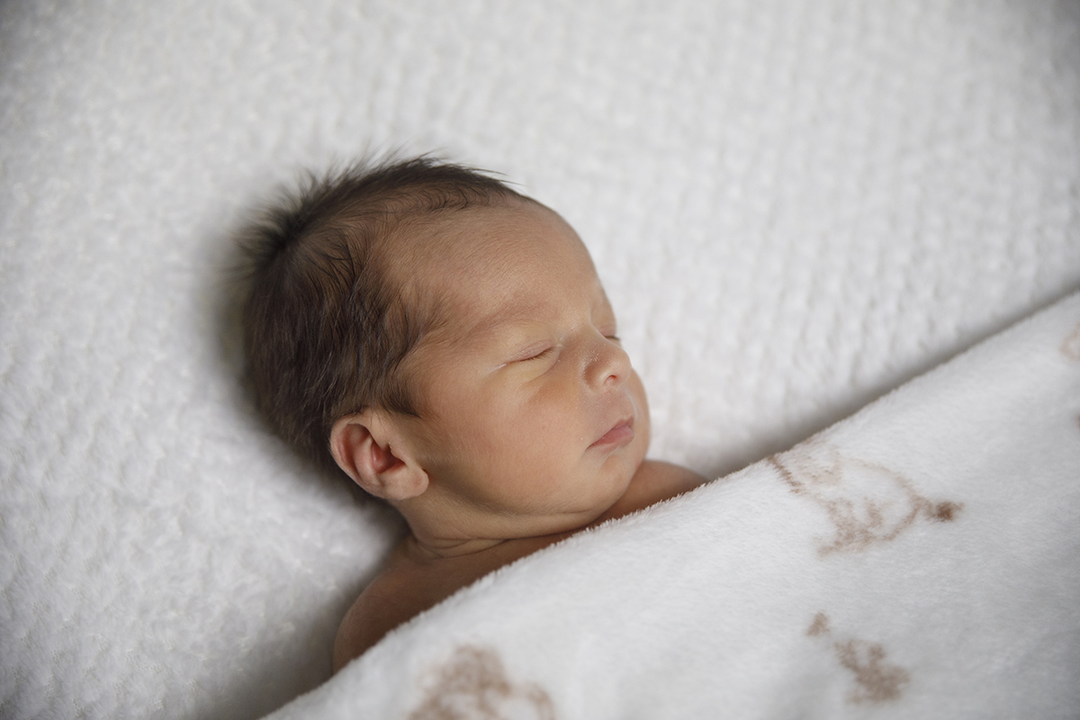 Gabriel está deitado, tapado até aos ombros com uma mantinha branca com desenhos castanhos simples. Dorme com a cabeça ligeiramente virada para o lado.