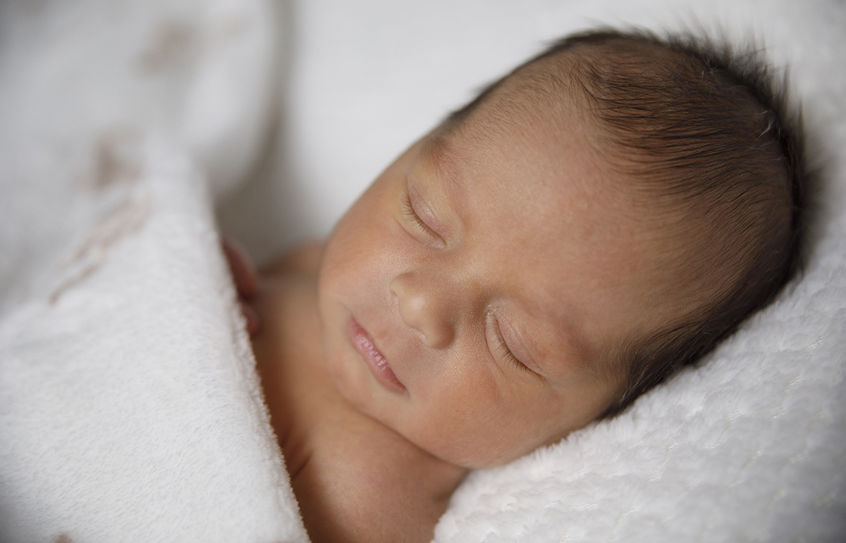 Um plano mais aproximado e de lado, focando em Gabriel a dormir com a mantinha até aos ombros. O fundo desfocado dá destaque ao bebé adormecido.