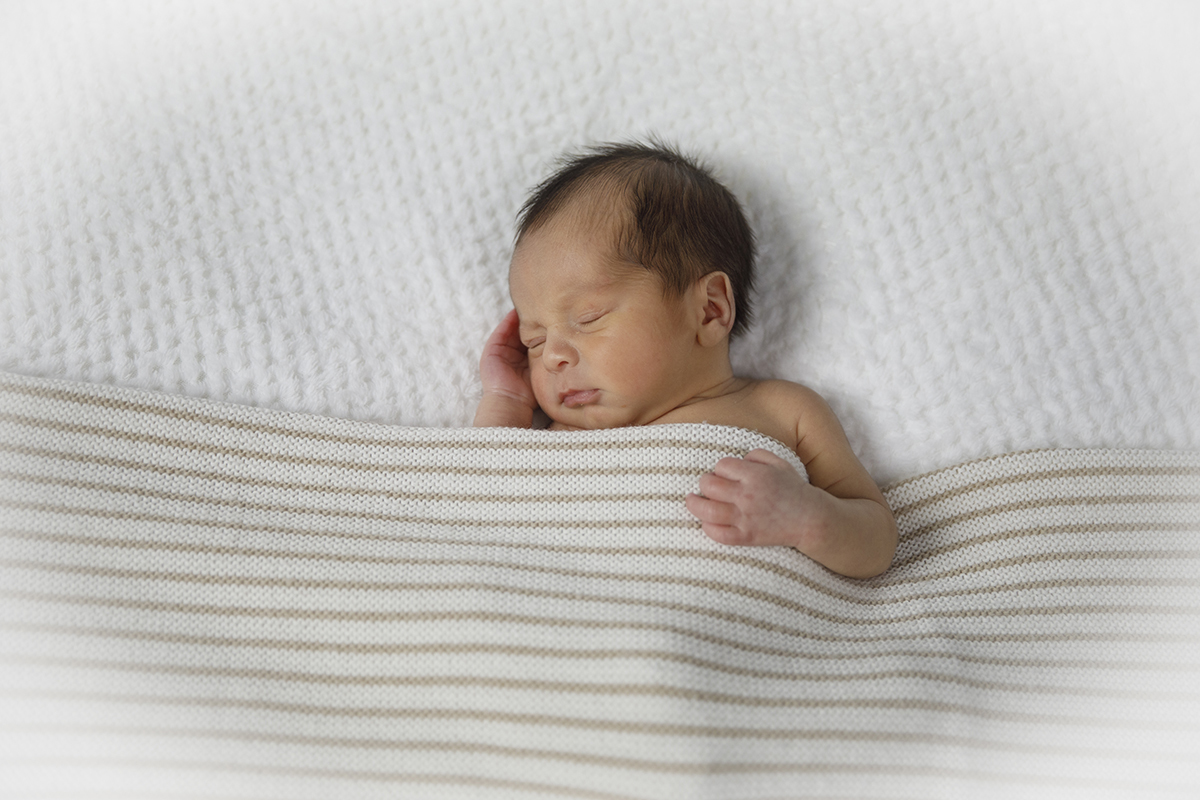 Um plano mais afastado onde Gabriel está no centro da fotografia, virado ligeiramente para o lado esquerdo. Está tapado até ao peito, com uma mãozinha sobre a manta e a outra junto ao rosto. A vinheta branca acrescenta suavidade à composição.