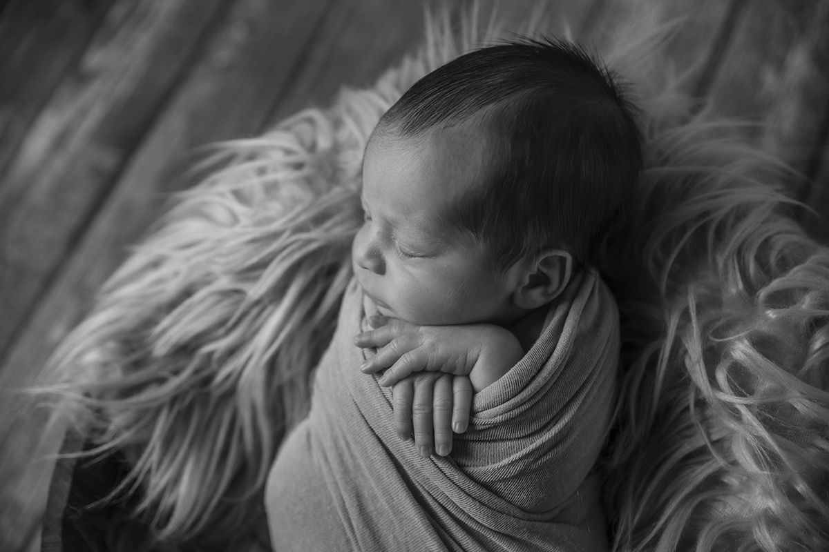 Ainda a preto e branco, Gabriel dorme numa caminha de pelo com o chão de madeira desfocado ao fundo; está enrolado numa manta, com a cabeça levemente virada e as mãos ao pescoço.
