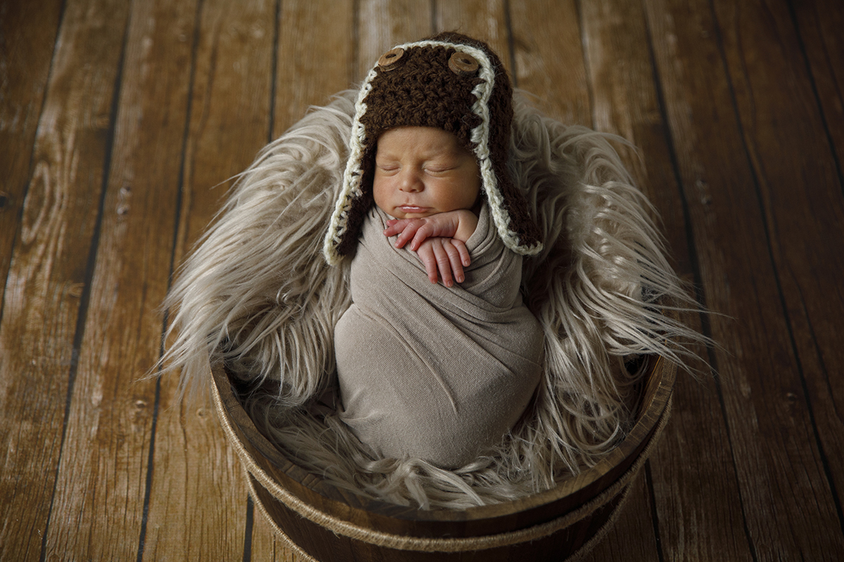 Agora a cores, vemos Gabriel deitado num círculo de madeira com manta felpuda, enrolado numa toalha bege, a dormir com as mãos ao lado do pescoço e um gorro de aviador castanho e branco.