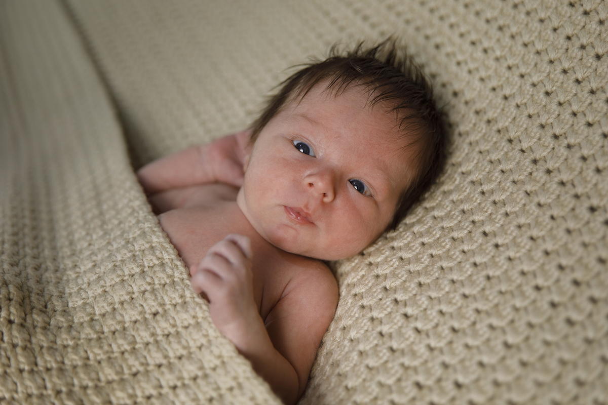 Lourenço está acordado, com os olhos entreabertos e uma expressão séria e curiosa. Uma mão ao pé da orelha e outra junto ao peito completam o enquadramento. O tecido claro que o envolve é delicado e parece feito à mão. Fotos recem nascido