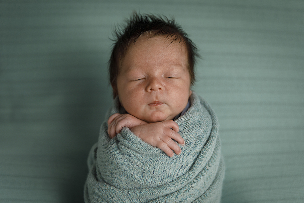 Lourenço surge envolto numa toalha verde-azulada, em perfeita harmonia com o fundo. Apenas a sua cabecinha e as pequenas mãos cruzadas estão visíveis, numa pose serena e adorável.