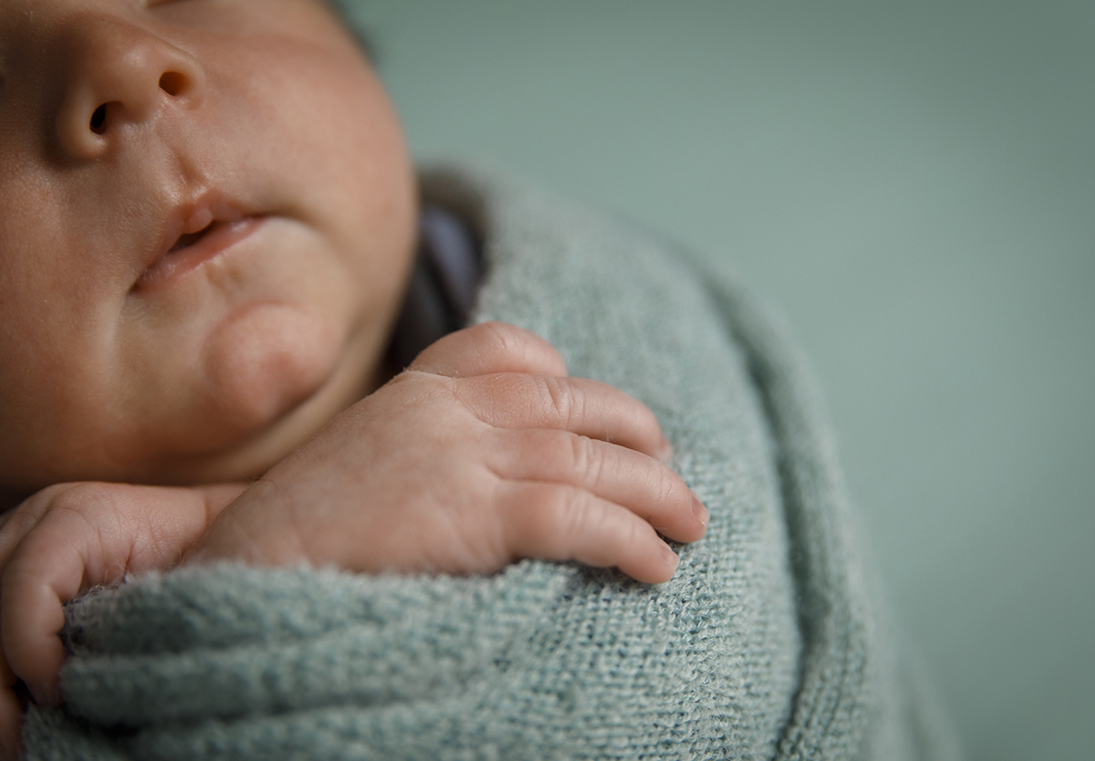 Um plano aproximado mostra as mãozinhas do bebé com maior detalhe, junto a parte do rosto delicado de Lourenço. O fundo, desfocado, reforça o foco na suavidade do momento. Sessao newborn