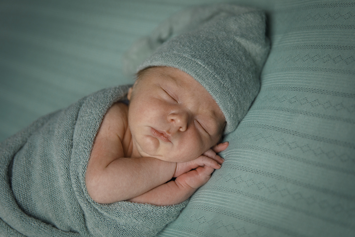 Tirada de cima ou de lado, esta imagem capta Lourenço com os braços fora da manta e as mãos suavemente contornando o pescoço e parte do rosto. Uma pose delicada e instintiva. Fotografia de newborn