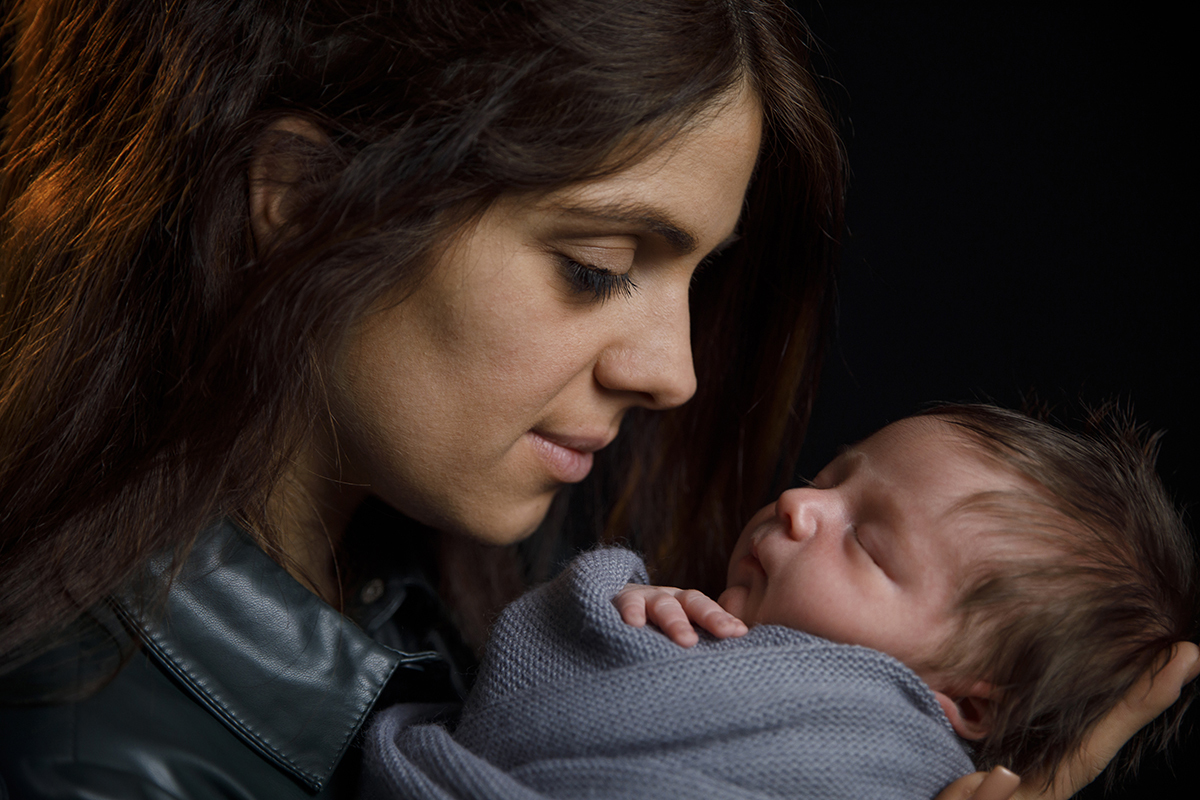 Plano aproximado dos ombros para cima, a mãe olha para o filho com seriedade e amor, enquanto Lourenço dorme profundamente nos seus braços. Uma imagem serena e poderosa.