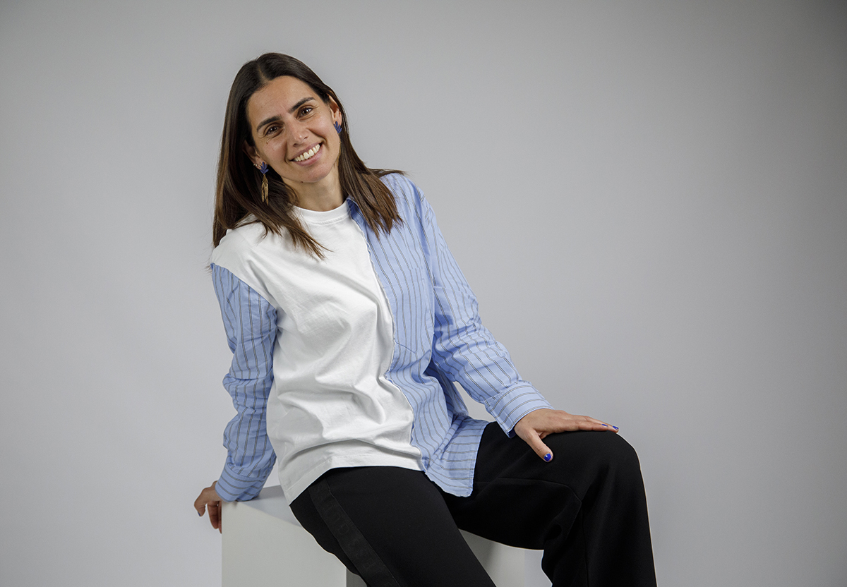 Contra um fundo branco, Sónia está sentada, com uma mão pousada num vértice do cubo maior e a outra sobre o joelho. Um plano dos joelhos para cima onde o sorriso feliz transmite simplicidade e bem-estar.