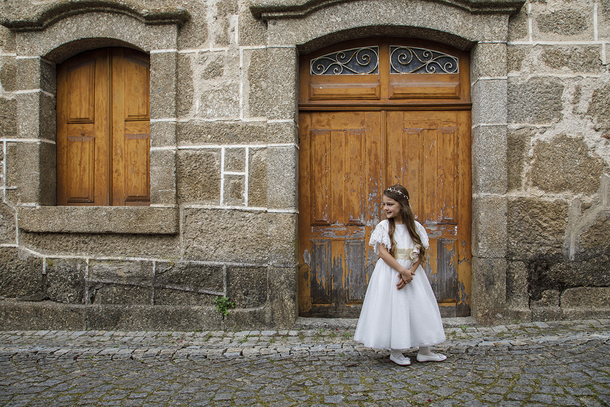 Letícia posa em frente à porta e janela semelhantes, num piso de paralelos. De frente, vira suavemente a cabeça, num gesto doce e expressivo.