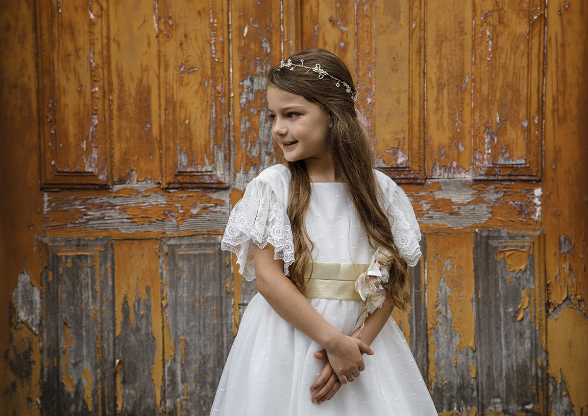 Junto a uma porta de madeira alaranjada e com tinta a descascar, Letícia está de frente, com as mãos sobrepostas e um sorriso sereno. O contraste da madeira rústica com a pureza do vestido cria um efeito visual envolvente.