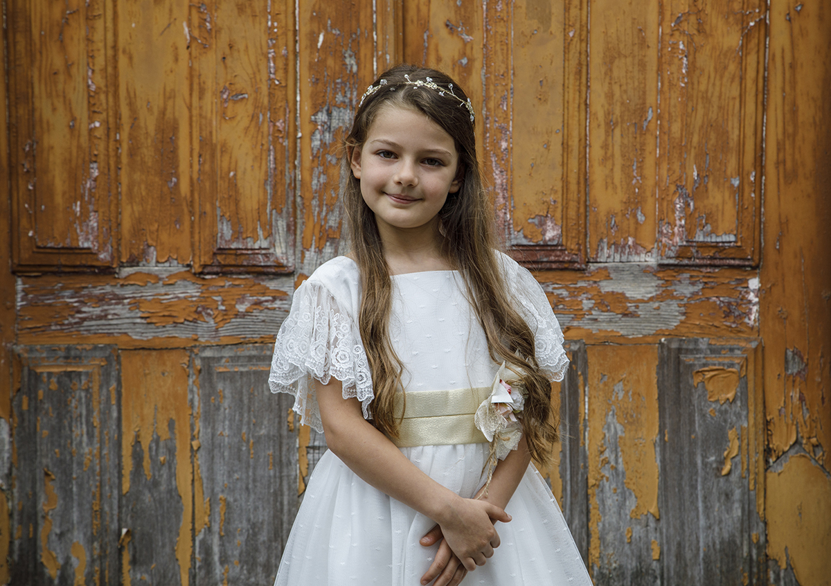 Num plano da cintura para cima, Letícia surge à frente da porta, com um sorriso leve e mãos sobrepostas. A serenidade da pose complementa o enquadramento.