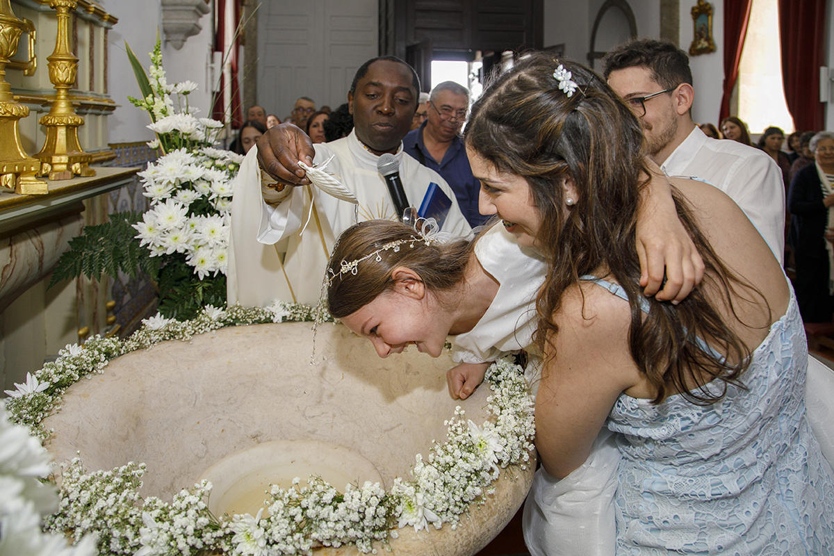 Chega o instante do Batismo. Letícia está debruçada sobre a Pia Batismal, apoiada por um casal, enquanto o padre derrama a água benta com a concha. A pia, decorada com flores brancas, torna o momento ainda mais especial. Fotografo Batizado