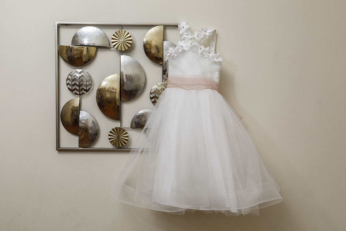 Contra uma parede branca e simples, destaca-se um quadro elegante em tons de dourado e prateado. Num dos cantos, pendurado com carinho, está o delicado vestido branco de batizado da Joana, com um doce pormenor rosa.