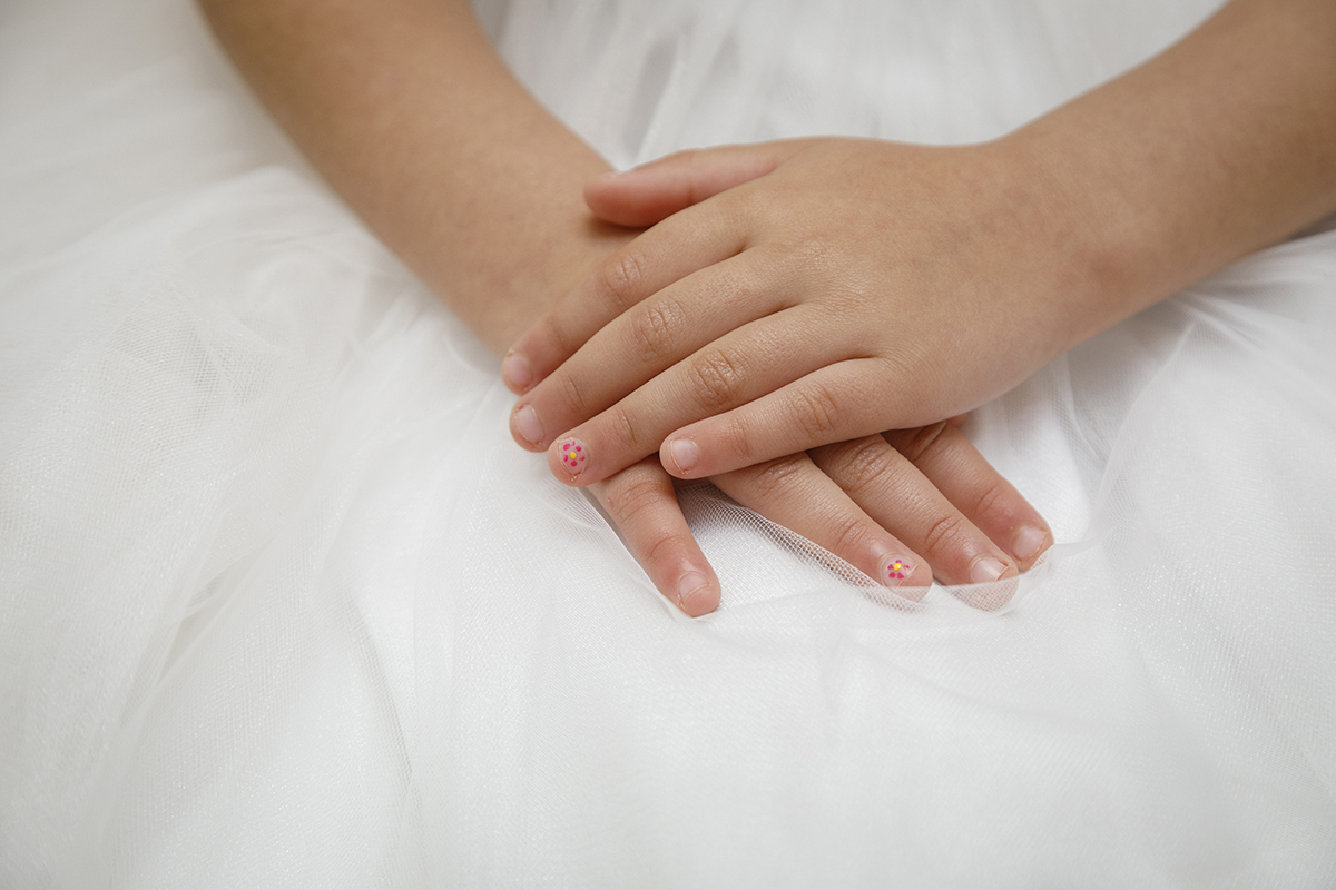Num plano mais aproximado, vemos o vestido com maior detalhe. As mãos da Joana repousam sobre ele com leveza, e um pequeno detalhe chama a atenção: nas unhas, uma florzinha rosa, que reflete a ternura do momento.