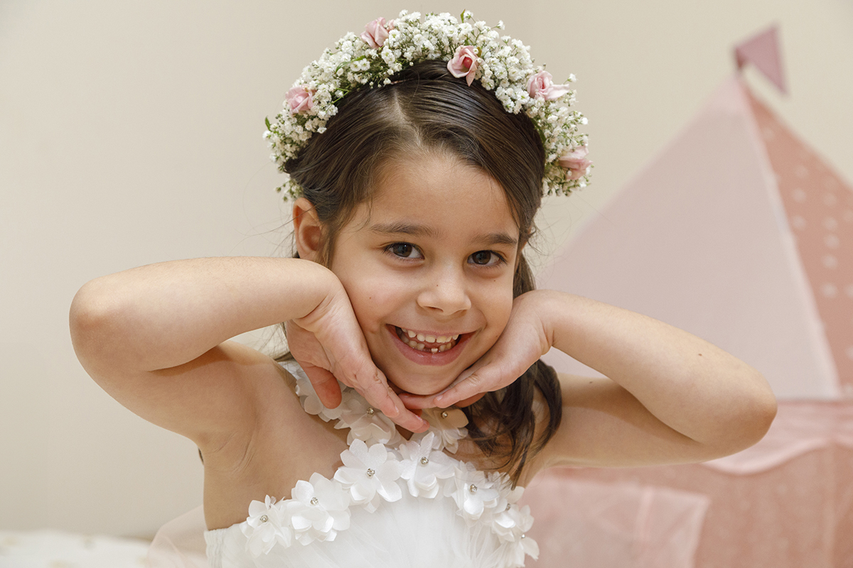 Joana sorri alegremente. As suas mãos contornam o rosto num gesto expressivo e doce. Ao fundo, um detalhe rosa, talvez uma tenda, completa esta pose encantadora. fotografia Batizado