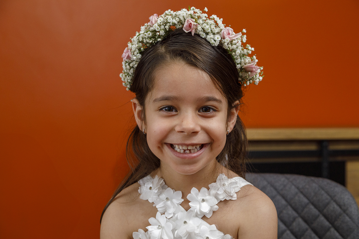 Num plano próximo, Joana mostra um sorriso largo e sincero. Usa uma coroa de flores brancas e rosas que combina perfeitamente com o vestido. As florzinhas rendadas ao pé do pescoço completam este look angelical.
