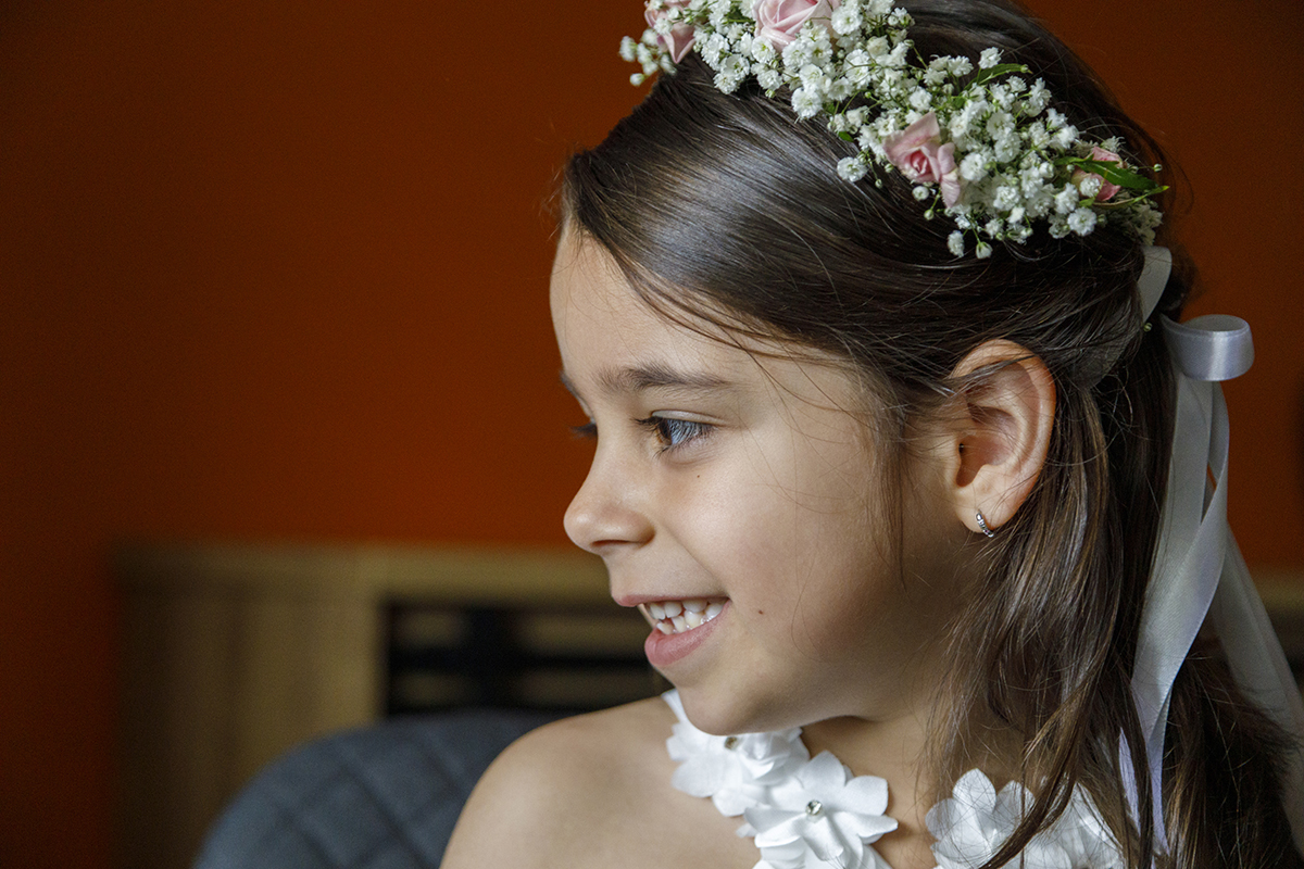 De perfil, Joana surge serena e feliz, com o laço branco no cabelo em destaque. O fundo desfocado foca toda a atenção na sua expressão. Reportagem Batizado