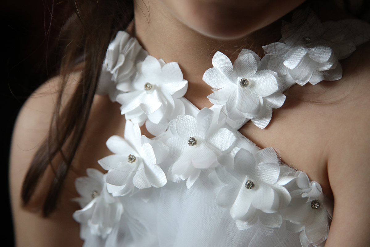 Um plano de detalhe foca na parte superior do vestido, flores rendadas brancas com bolinhas prateadas ao centro, mostrando a riqueza do tecido e o cuidado nos pormenores.