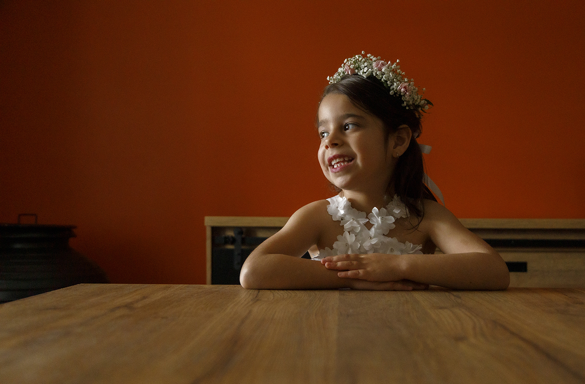 Sentada, com os braços pousados sobre uma mesa de madeira, Joana olha para o lado sorridente. Uma luz suave ilumina o seu rosto, vindo, provavelmente, de uma janela próxima.