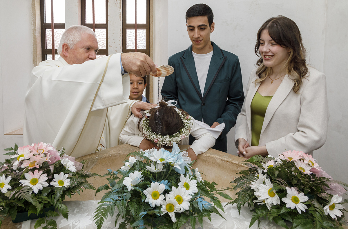 O momento central do dia. Joana está debruçada sobre a pia batismal, enquanto o padre derrama a água benta sobre a sua cabeça. Ao lado, um casal e uma criança assistem com carinho.