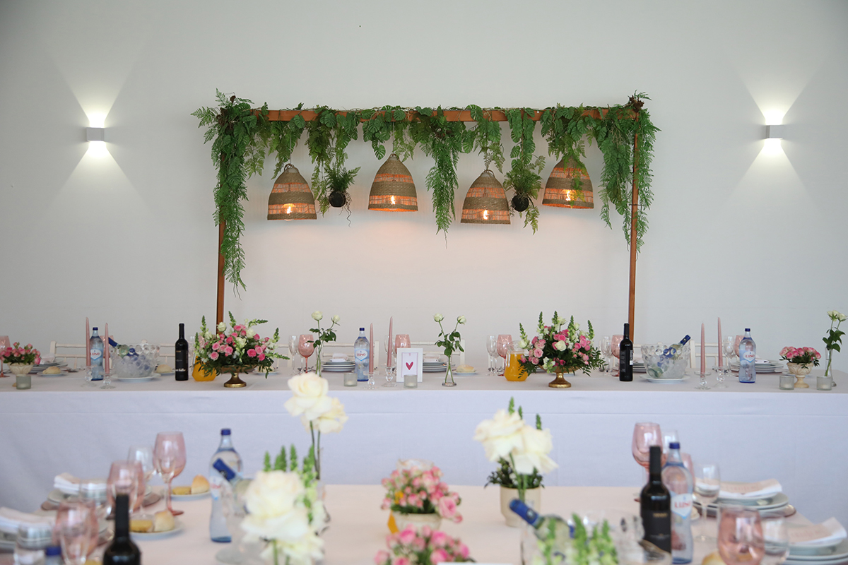 Num ambiente acolhedor, vemos uma parede branca com dois candeeiros acesos e mesas decoradas com flores e copos cor-de-rosa, tudo cuidado ao pormenor.