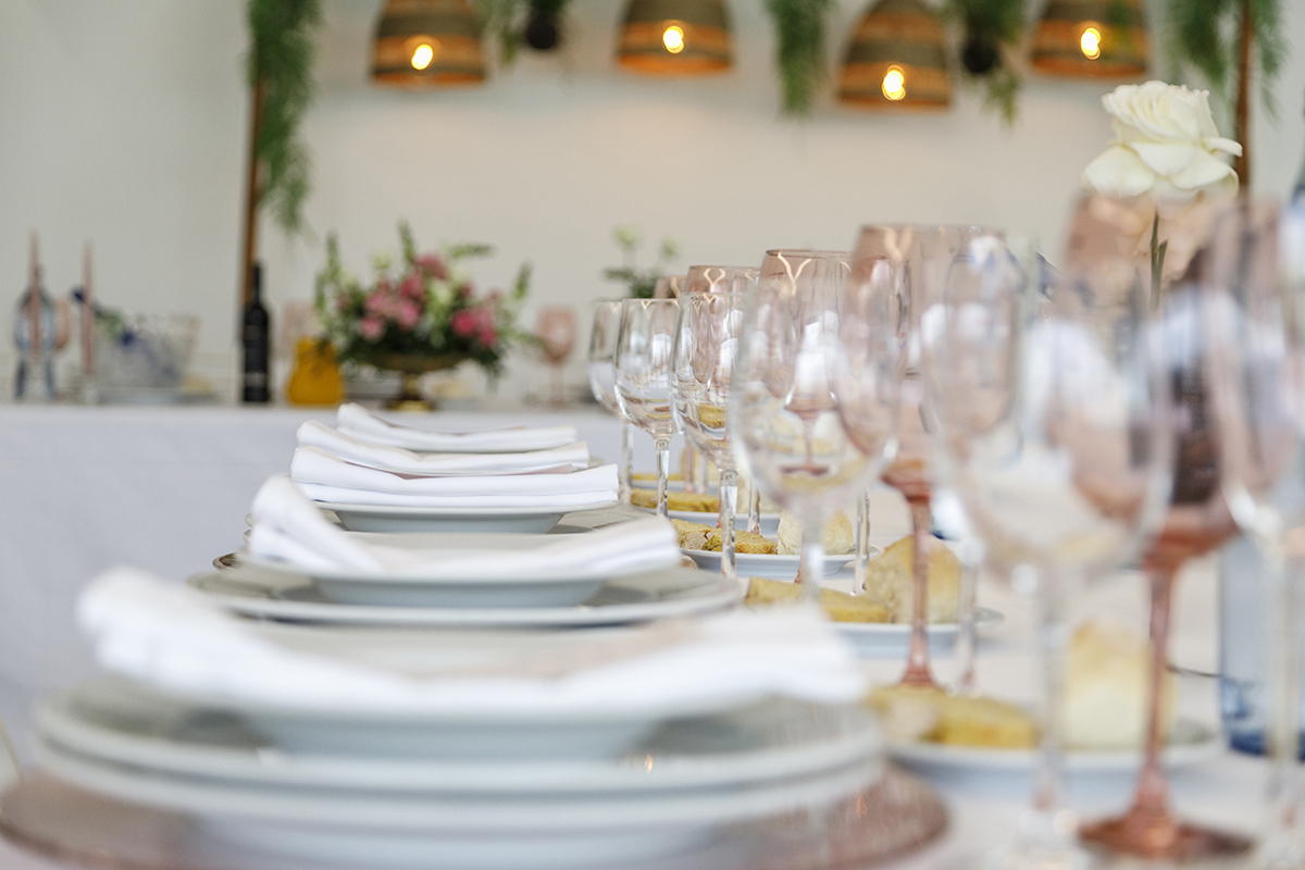 Um plano mais próximo mostra a delicadeza da receção, três pratos, um guardanapo elegante e copos, um deles de cristal transparente e outro rosa.
