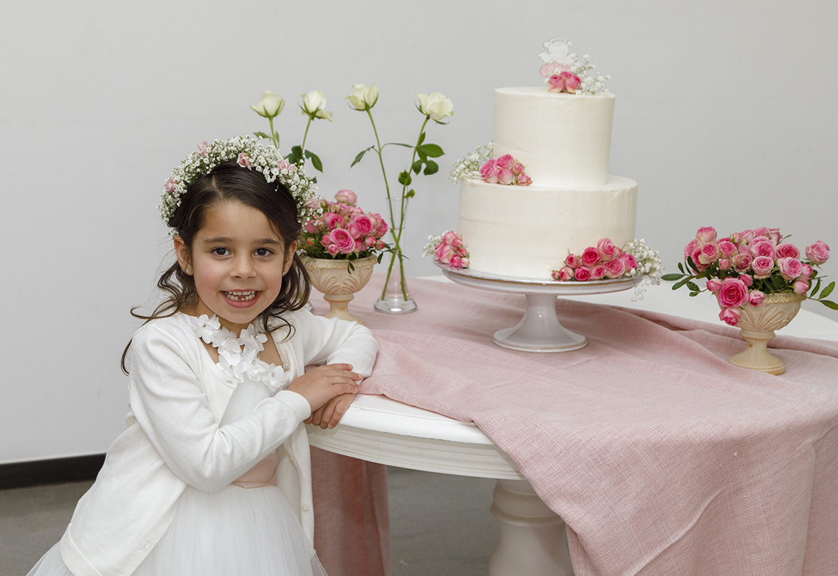 Joana está encostada à mesa, ao lado do seu lindo bolo branco decorado com flores rosas, com um sorriso de pura felicidade.