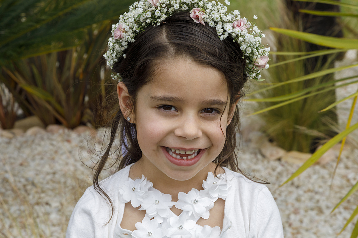 Com um fundo de plantas e pedras brancas desfocado, Joana sorri radiante, a sua coroa de flores e o pormenor do vestido junto ao pescoço completam o cenário com graciosidade.
