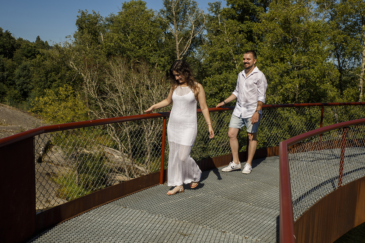 Sandra e Rafael caminham lado a lado pela ponte, cada um com a mão no varão, envoltos pela paisagem outonal banhada por um sol intenso que aquece o cenário e ilumina os seus passos.