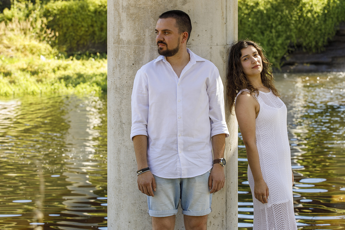 Junto ao pilar, Rafael olha para o lado, Sandra encara a lente com leveza.