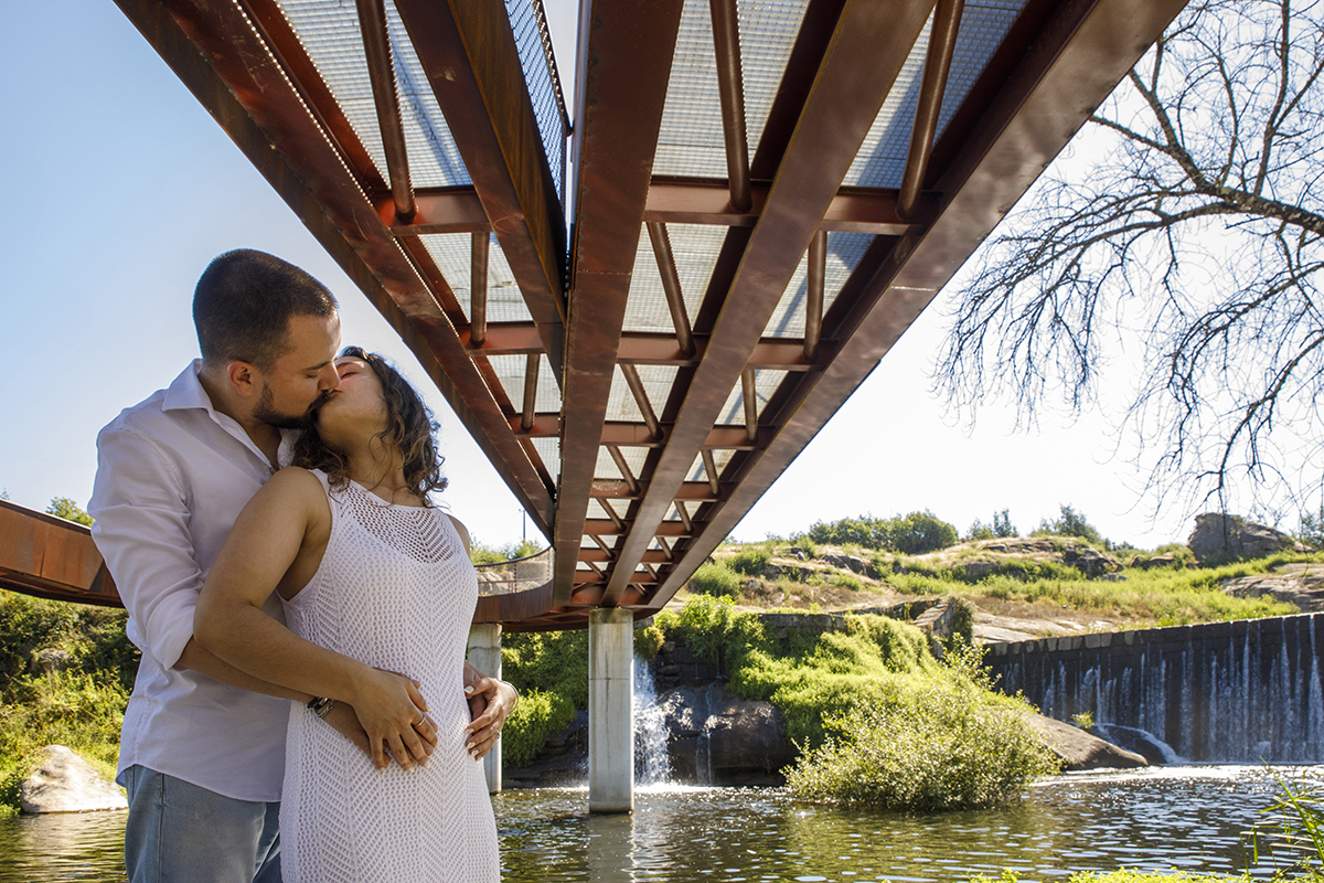 Debaixo da ponte, abraçados, trocam um beijo. A paisagem natural completa o quadro.