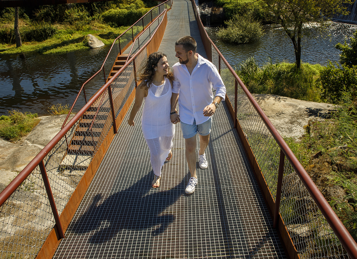 Num ângulo picado, caminham sobre a ponte, olhos nos olhos, partilhando sorrisos.