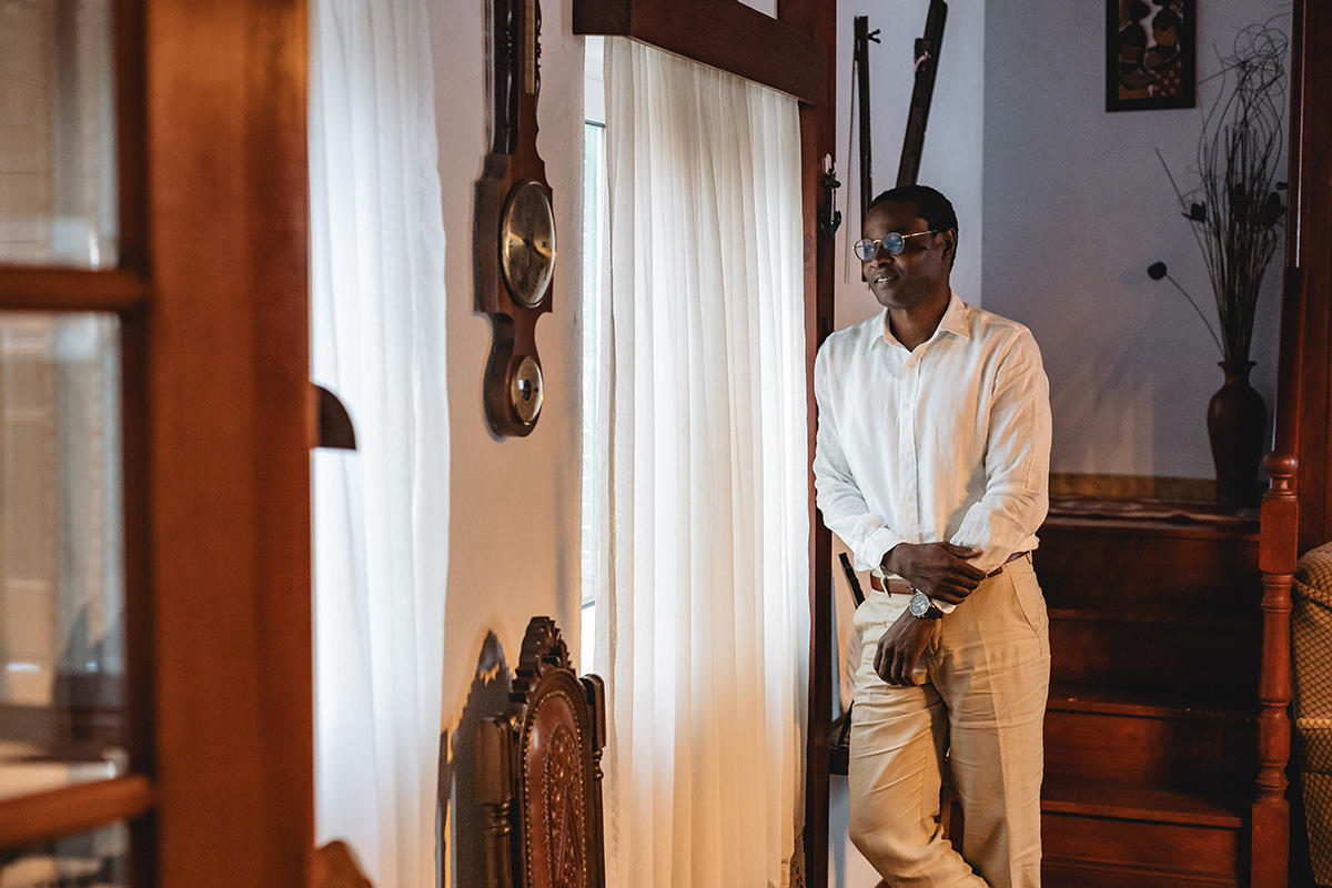 De camisa branca e calças de linho claro, ele encosta-se á janela, sereno, entre luz e sombra. O ambiente acolhedor da casa e os detalhes de madeira reforçam a intimidade do momento.