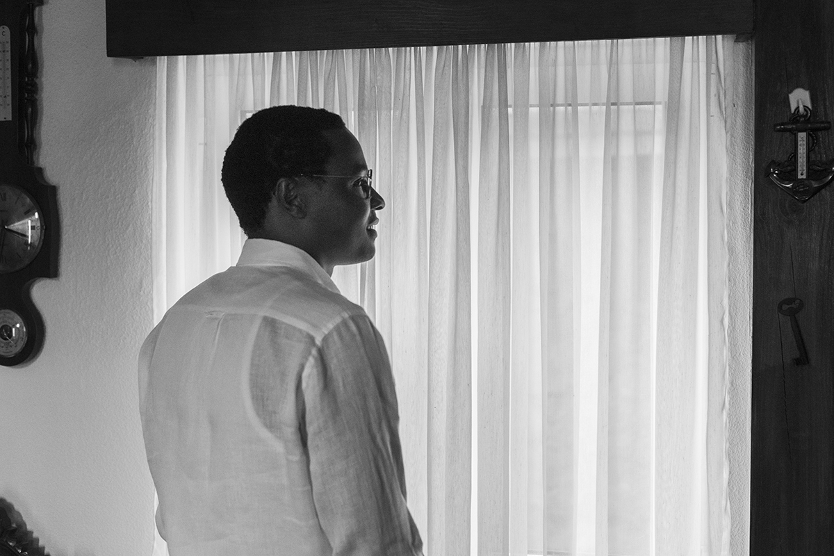 A preto e branco, com o olhar voltado para a janela, ele deixa-se envolver pela luz que atravessa a cortina. O silêncio da cena transmite uma paz profunda, onde cada detalhe respira emoção.