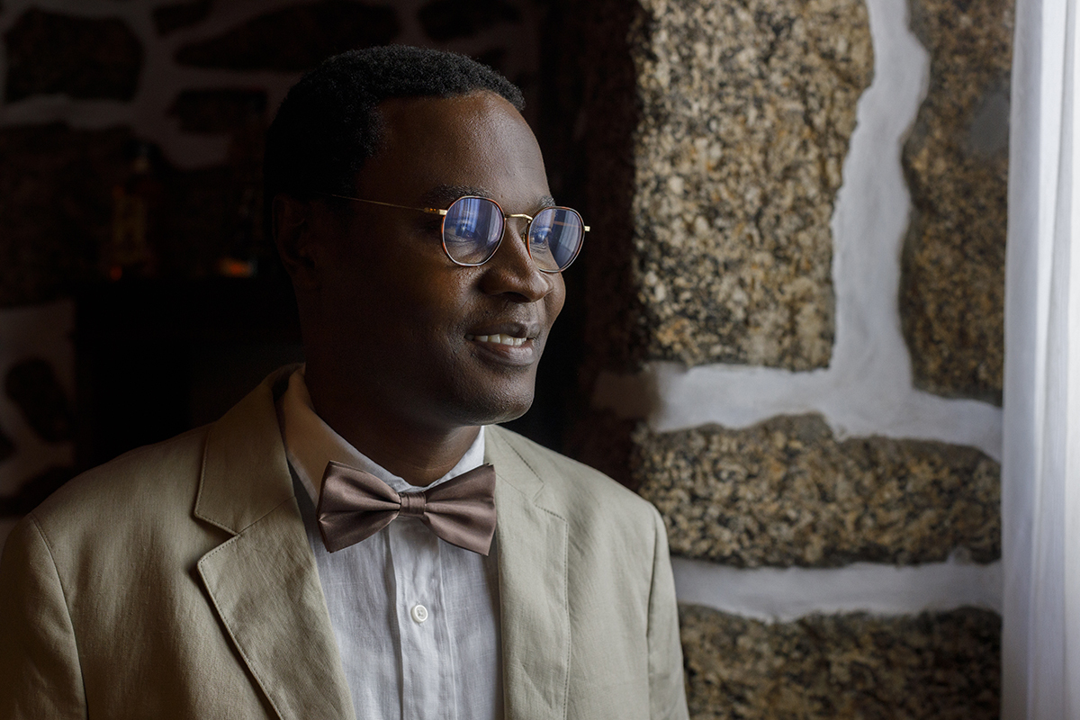 Num enquadramento de luz natural, Hélder sorri com tranquilidade junto a uma parede de pedra, com o olhar pousado num ponto longínquo. O instante revela calma e antecipação.