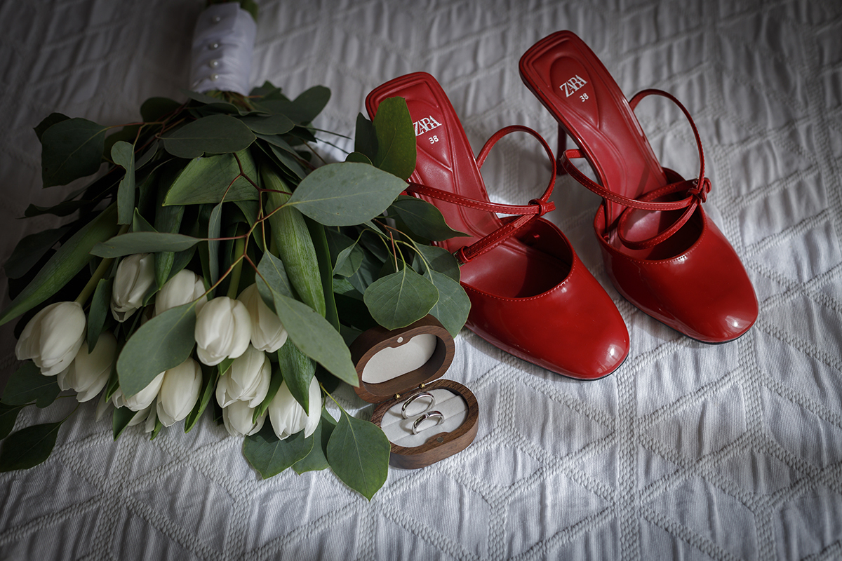 Um registo artístico dos acessórios, onde o vermelho vibrante dos sapatos contrasta com a delicadeza do ramo de flores. Ao centro, as alianças repousam discretamente, simbolizando o compromisso eterno. Fotografia casamento