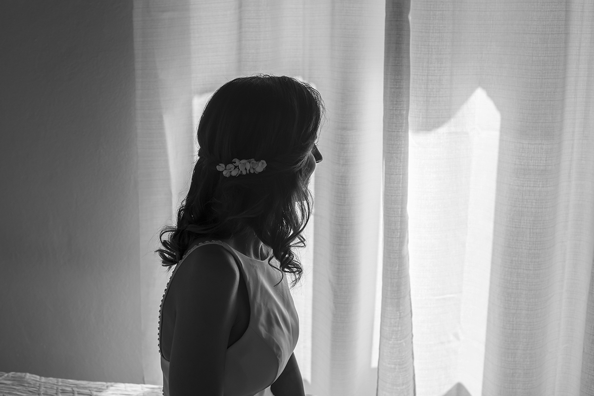 Ju contempla o dia pela janela, envolta em luz suave. A fotografia, a preto e branco, eterniza esse instante de silêncio e reflexão.