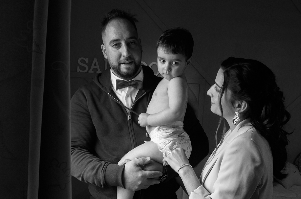 Num gesto de afeto, a mãe toca suavemente na perna do filho, enquanto o pai o segura com carinho ao colo. Os olhares da mãe e do pequeno Salvador cruzam-se num instante sério, mas repleto de amor silencioso.