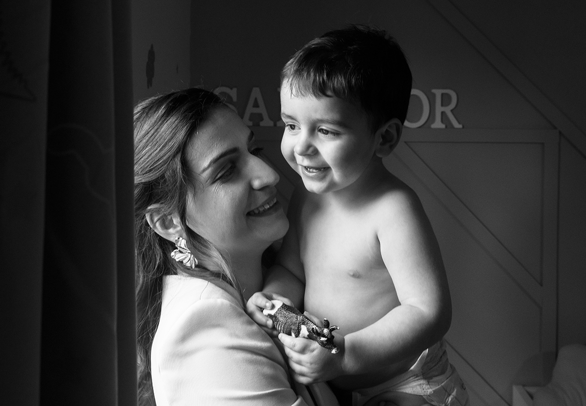 Ao colo da mãe, Salvador observa o mundo pela janela com um brilho nos olhos. Ela, com um sorriso tranquilo, admira-o como quem vê um milagre.