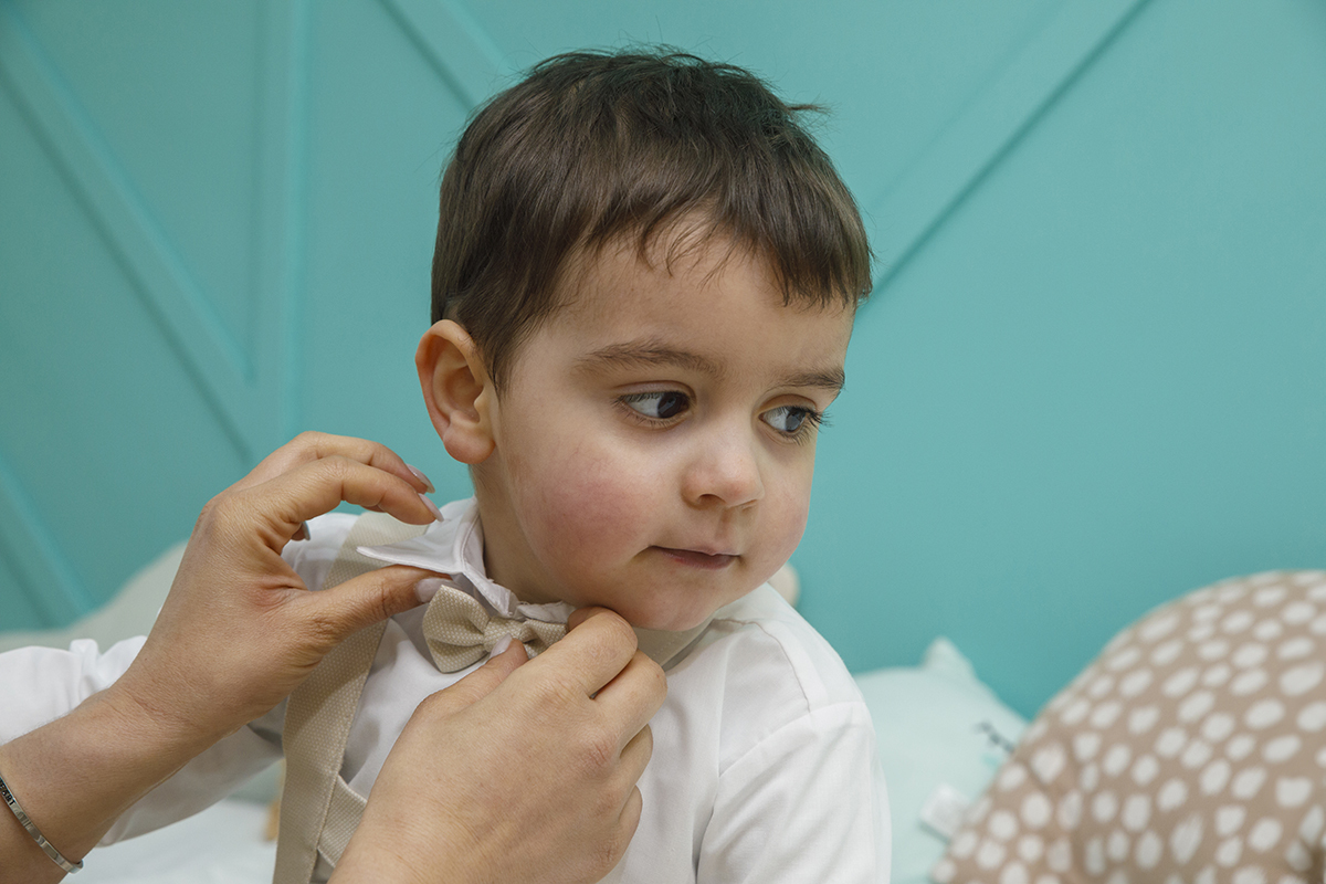 Com um olhar atento, Salvador fixa-se num ponto enquanto a mãe lhe compõe o laço. Um instante de cuidado e cumplicidade. Reportagem Batizado