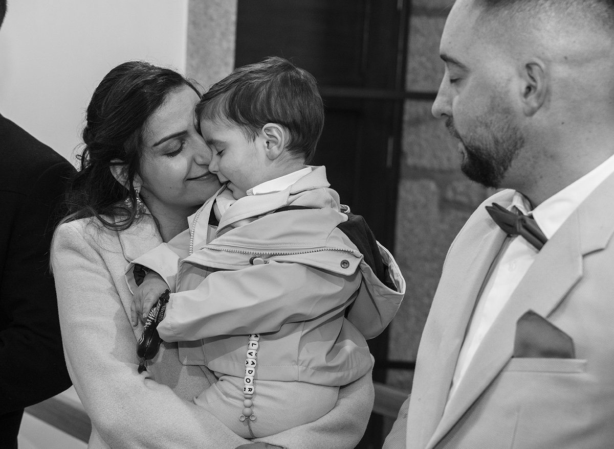 Ao colo da mãe, Salvador encosta a carinha à dela. Ambos trocam um sorriso tranquilo, enquanto o pai observa, sereno, ao fundo.