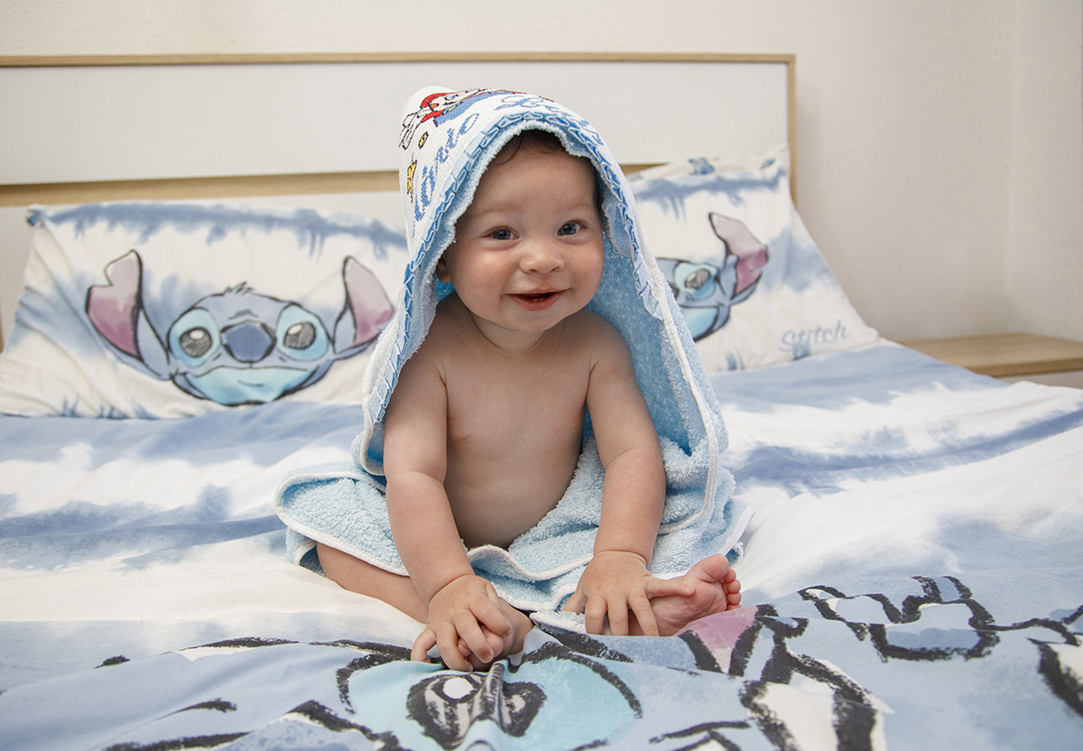 Sentado em cima da cama, Mário está enrolado na sua toalha, com as mãos sobre os pés e um sorriso bonito e contagiante.Fotografia pela lente do fotografo Ricardo Coimbra