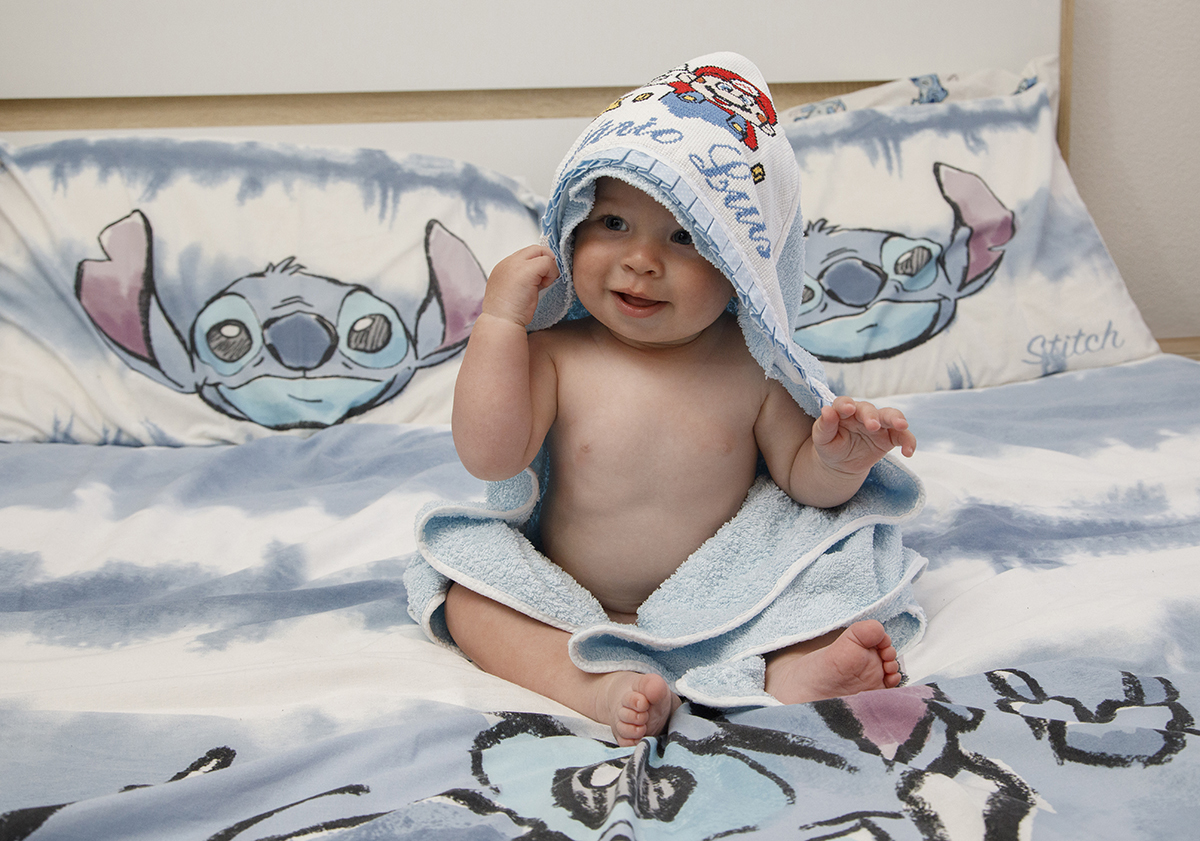 Enrolado na sua toalha azul bebé, Mário senta-se na cama, olhando para o lado esquerdo, com um pequeno sorriso no rosto.