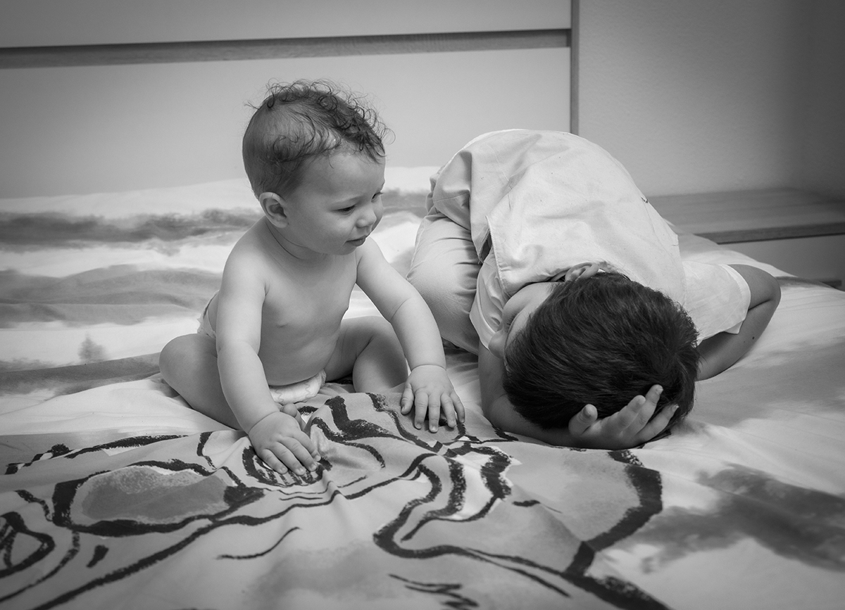 Sentado na cama, o pequeno Mário olha para o seu irmão, que tem a cabeça apoiada na cama, partilhando um olhar terno que leva a um sorriso no rosto do Mário.