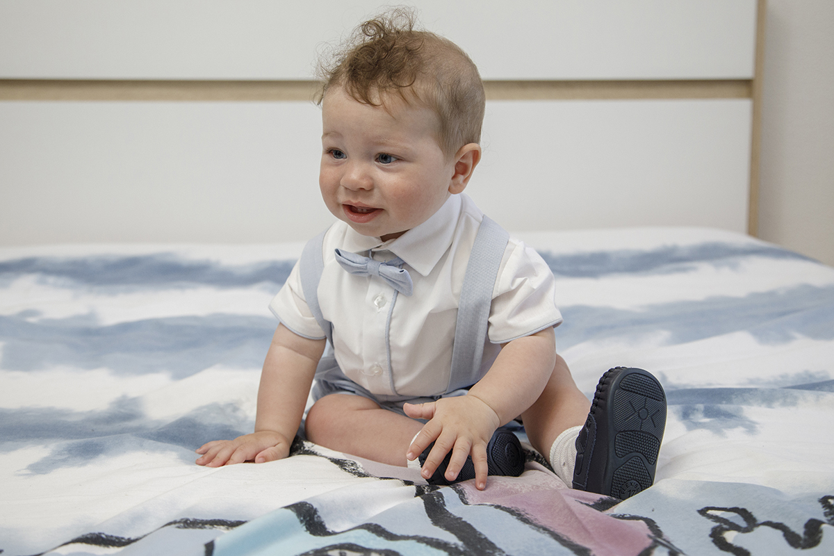 O pequeno Mário está sentado na cama, com uma mão pousada na colcha e a outra sobre o seu pé, com uma expressão fofa e serena.