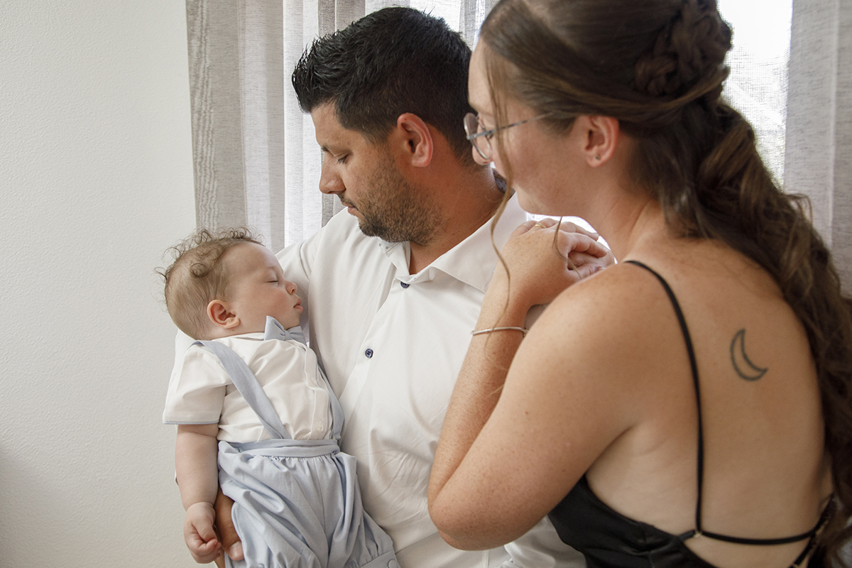 O pai segura o pequeno Mário ao colo, com a mãe ao seu lado. Ambos os pais olham com ternura para o filho adormecido, num momento de pura contemplação.