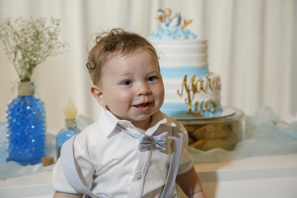 Com um olhar atento e um sorriso a surgir, Mário posa junto à sua mesa decorada, o bolo bonito ao atrás promete doces momentos.