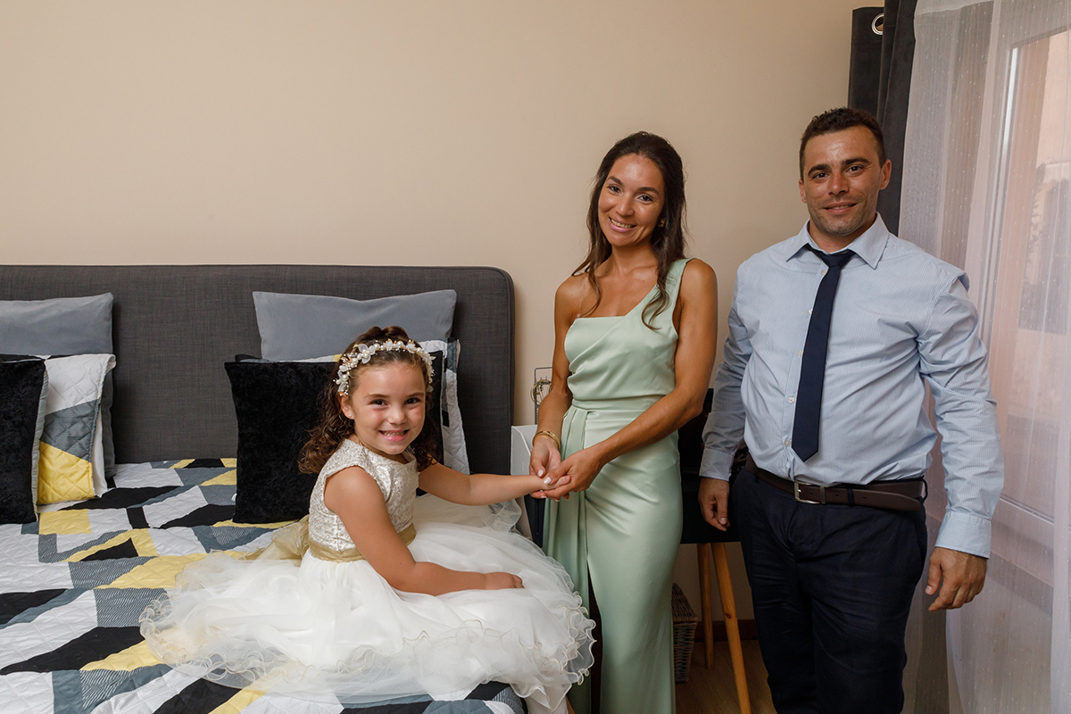 Entre sorrisos cúmplices, Iris pousa uma mão sobre o vestido e outra nas mãos da madrinha, com o padrinho ao lado, num momento cheio de ternura.